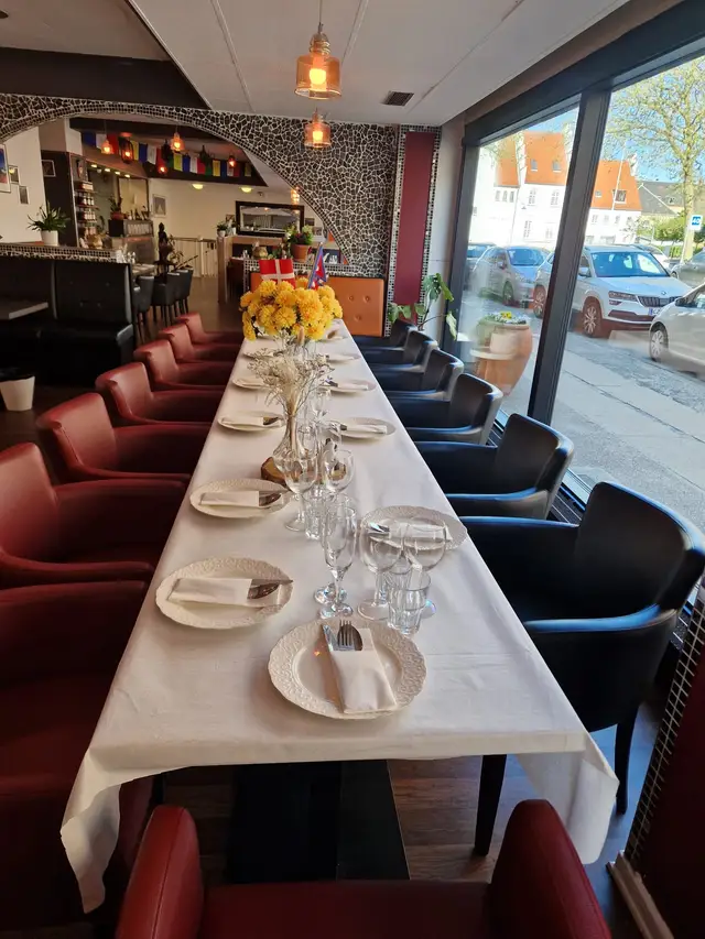 A long white-clothed table with plates, glasses and yellow chrysanthemums, red leather armchairs down one side and daylight from the street window