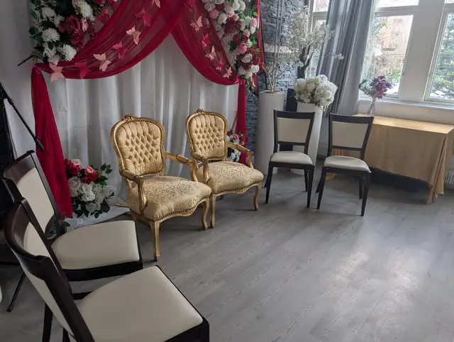 Two gilded armchairs on a white draped backdrop with crimson swags, pink butterflies and roses, beside daylit windows