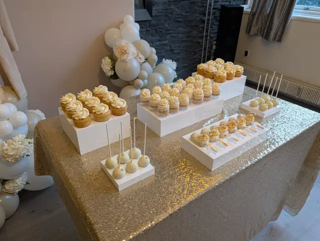 Dessert table on a gold sequin cloth: vanilla cupcakes and white cake pops on white risers beside a white balloon and hydrangea garland