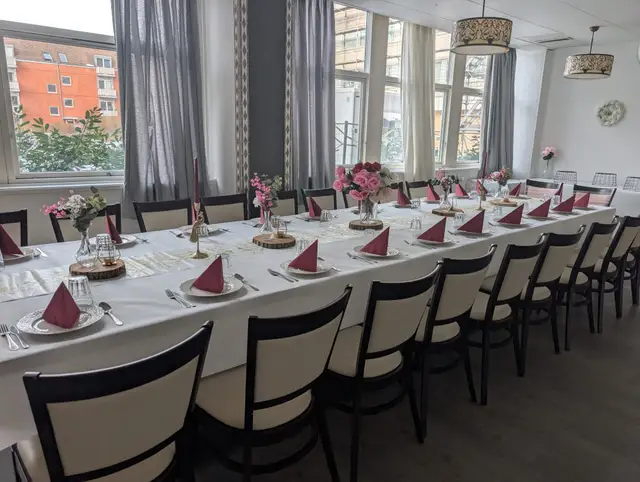 A long table beside the hall's tall curtained windows, set with raspberry napkins, pink roses and wood slices in daylight